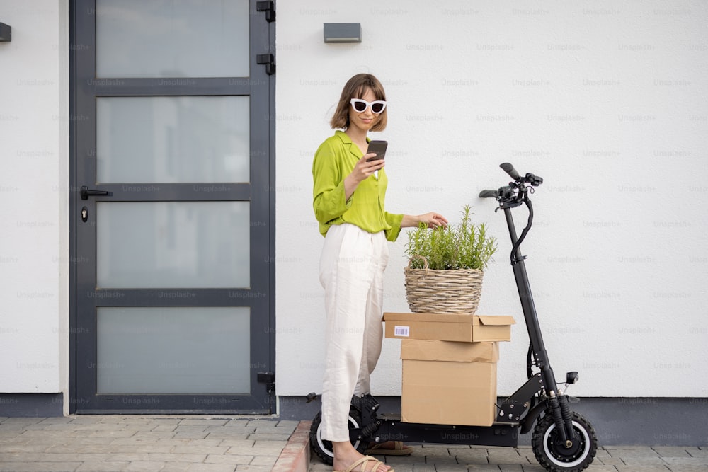 Femme utilisant un téléphone debout sur un porche de sa maison avec un scooter électrique et des colis à proximité. Concept de durabilité et de mode de vie moderne respectueux de l’environnement