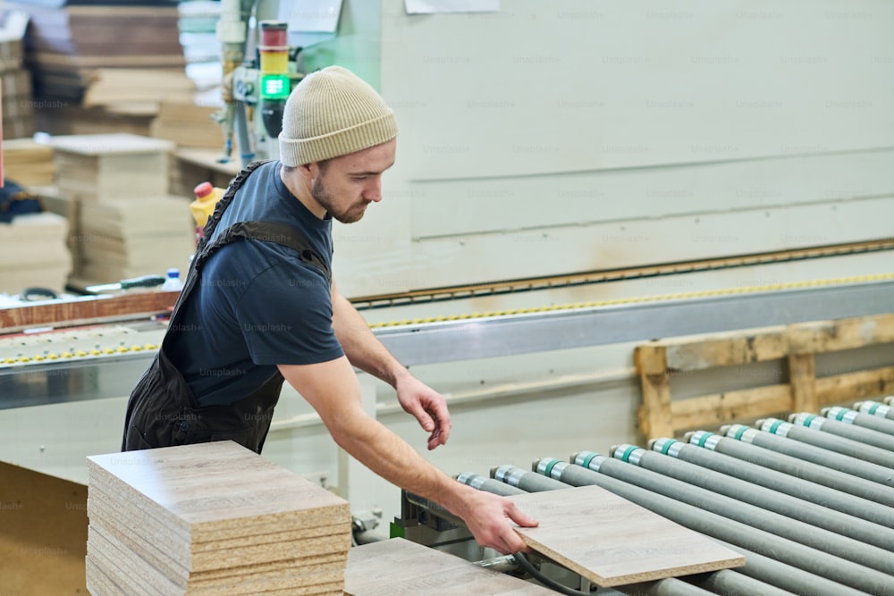 Young carpenter in overall putting wooden boards on machine during production of furniture at factory