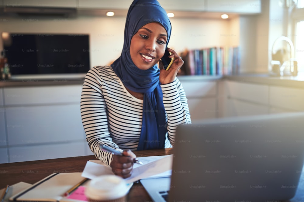 ヒジャブをかぶった若いアラブ人女性起業家がキッチンのテーブルに座り、自宅で仕事をしながら携帯電話で話し、ノートパソコンを使う