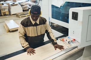 Artesano africano con auriculares protectores y uniforme concentrado en su trabajo de carpintería en la máquina