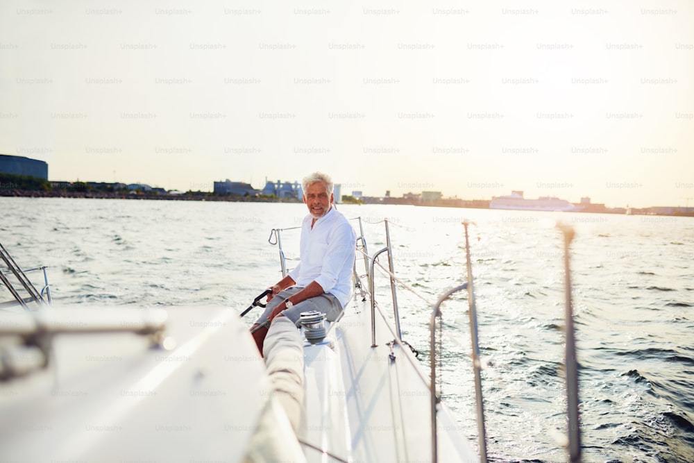 Reifer Mann sitzt allein auf dem Deck eines Bootes, das an einem sonnigen Tag entlang der Küste segelt