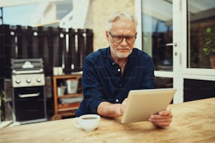 Homem idoso sorridente sentado em uma mesa do lado de fora em seu pátio trabalhando on-line com um tablet digital e bebendo uma xícara de café