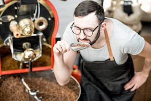 Hombre comprobando la calidad de los granos de café de pie con una cuchara cerca de la máquina tostadora en la tostadora