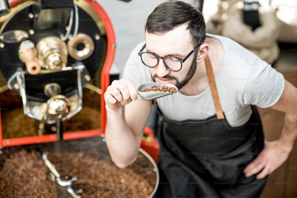 Homme vérifiant la qualité des grains de café debout avec une cuillère près de la machine de torréfaction à la torréfaction
