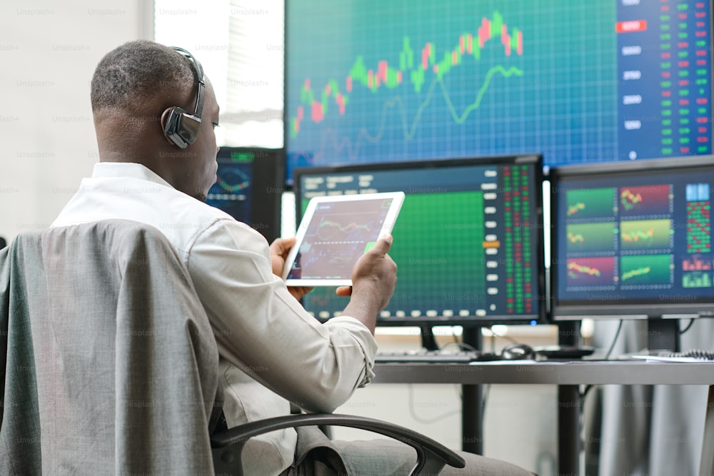 Giovane uomo nero che tiene la tavoletta digitale che lavora con le statistiche di trading azionario che guarda i grafici e i grafici della valuta