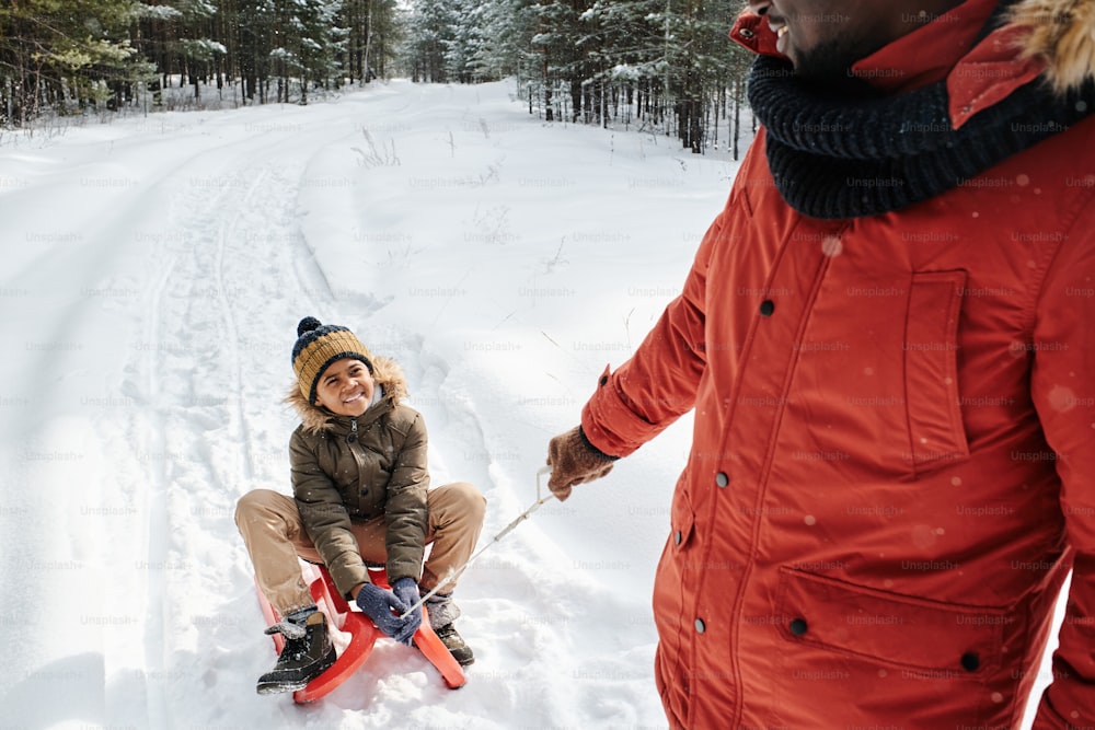 Niño lindo feliz en ropa de invierno mirando a su padre tirando del trineo con él mientras se mueve por la carretera en medio de bosques profundos