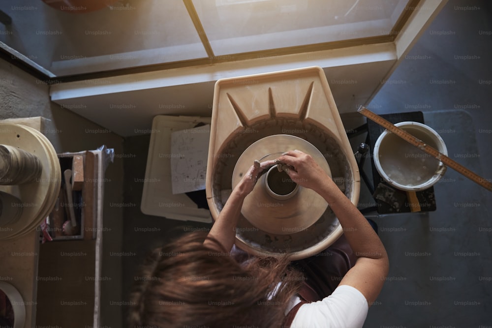 Artigiana seduta nel suo laboratorio di ceramica che usa le mani per modellare un pezzo di argilla bagnato che gira su un tornio di ceramica