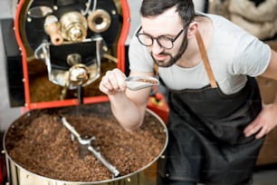 Homme vérifiant la qualité des grains de café debout avec une cuillère près de la machine de torréfaction à la torréfaction