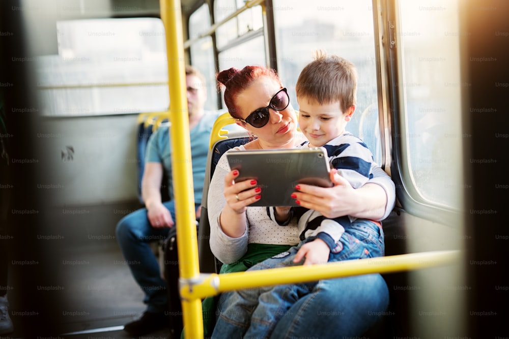 Una señora madura sostiene a su pequeño niño pequeño en su regazo y le muestra videos de tableta mientras viaja en autobús.