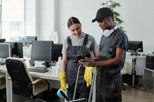 Young African American male cleaner showing female colleague his playlist in smartphone while standing in large openspace office