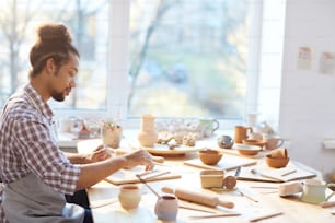 Side view of young mixed race male potter making beautiful clay souvenirs while sitting at table in workshop