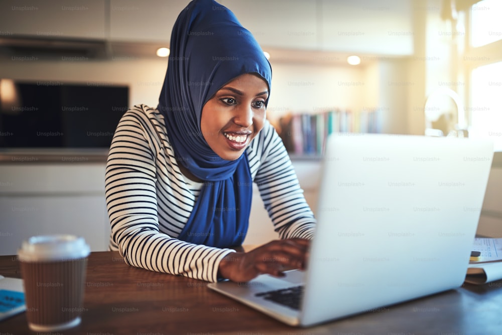 Lächelnde junge arabische Frau mit Hijab sitzt an ihrem Küchentisch und benutzt einen Laptop, während sie von zu Hause aus arbeitet
