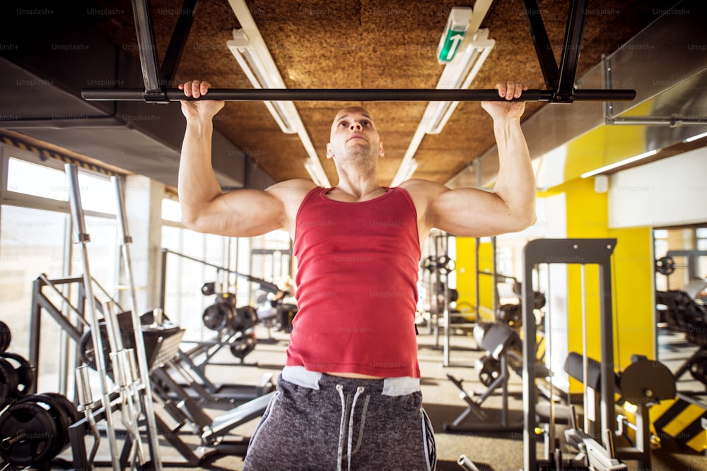 Close up front view of motivated and focused strong muscular active healthy young bald man working pull ups in the modern gym.