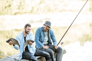 朝の湖で釣りをしているときに木製の桟橋に座って釣った魚を手にした2人の幸せな漁師