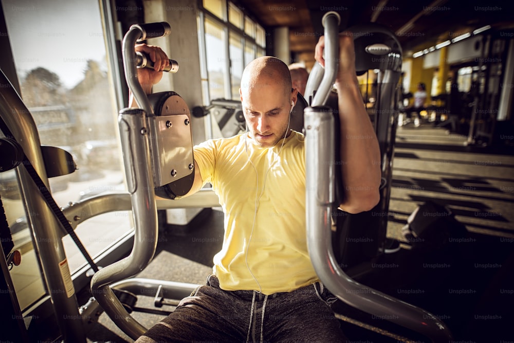 Portrait of strong muscular active healthy young bald man doing chest exercises on the machine and having earphones.