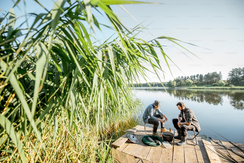 Two fishermen relaxing during the picnic on the beautiful wooden pier with green reed on the lake in the morning