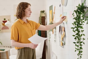 Jeune femme d’affaires prospère avec un document debout devant le mur avec un ensemble de photos et des papiers à lettres collants sous l’horloge