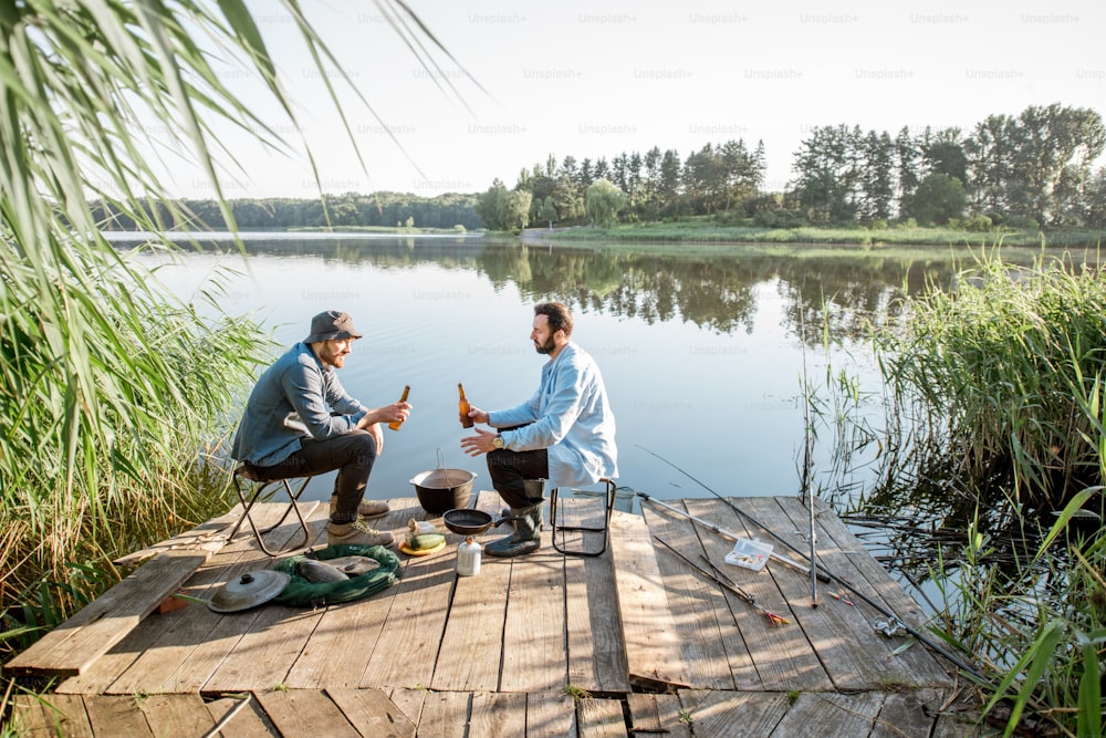 Vista del paisaje en el lago con dos amigos masculinos sentados juntos con cerveza durante el proceso de pesca