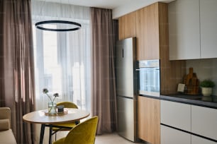 Mesa de cocina redonda y dos cómodas sillas de color amarillo mostaza de pie entre ventana con cortinas pastel y refrigerador