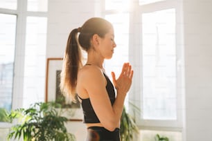 Young fit woman practice yoga doing asana namaste in light yoga studio with green house plant