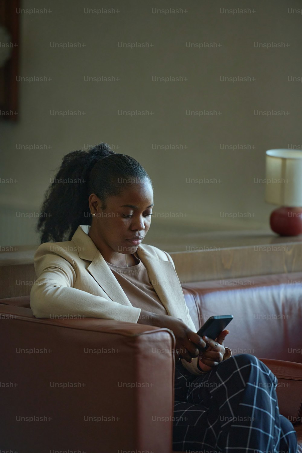 Young elegant black woman in formalwear texting in smartphone or communicating in video chat while relaxing on sofa in hotel room