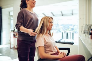 Lächelnde junge blonde Frau sitzt in einem Salonsessel und diskutiert ihre Haare mit ihrem Friseur