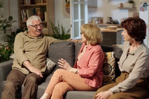 Group of aged friendly people in casualwear discussing latest news while sitting on soft couch in living room during gathering