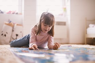 Hermosa y encantadora niña pequeña acostada en la alfombra de su habitación y jugando con un gran mapa.