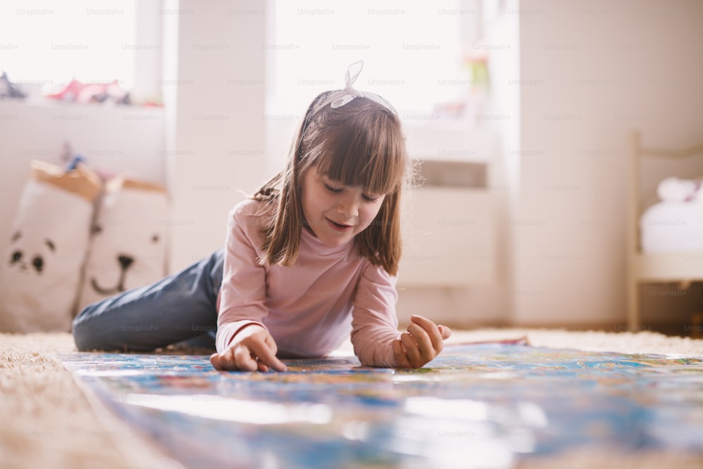Bella bambina affascinante sdraiata sul tappeto della sua stanza e che gioca con una grande mappa.