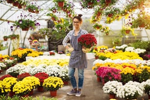 Hermosa florista joven sonriente con anteojos de pie en el invernadero lleno de flores mientras sostiene la maceta y mira a la cámara.