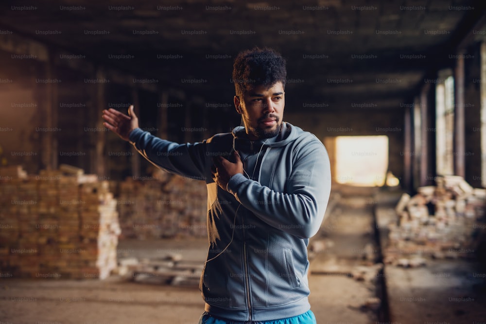 Close up portrait of active afro-american young attractive athletic man doing full hand stretching workout inside of the abandoned place.