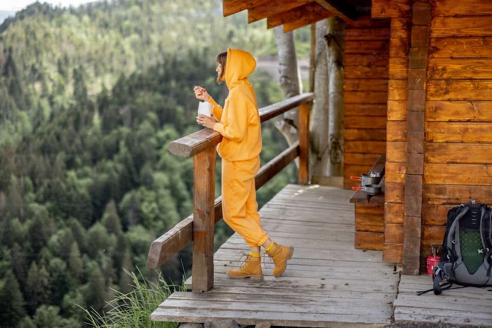 A jovem mulher desfruta de uma grande paisagem montanhosa e come comida sublimada para caminhadas, enquanto está em pé em um terraço de madeira de casa minúscula. Conceito de solidão na natureza e comida para caminhadas