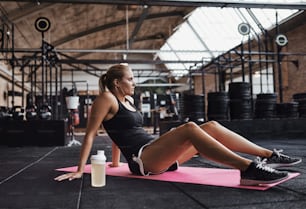 Giovane donna bionda in forma in abbigliamento sportivo seduta su un tappetino per esercizi sul pavimento di una palestra mentre si prende una pausa da un allenamento