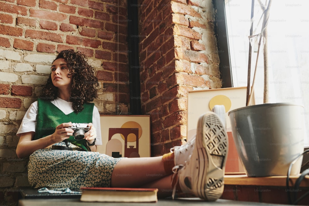 Young woman in casualwear holding retro photocamera while sitting against red brick wall by window in loft apartment and having rest