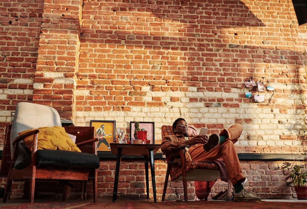 Young relaxed African American man sitting in armchair by brick wall and scrolling in mobile phone or communicating in video chat