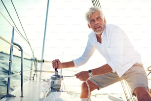 Hombre maduro de pie en la cubierta de un barco trabajando un cabrestante mientras sale a navegar en una tarde soleada