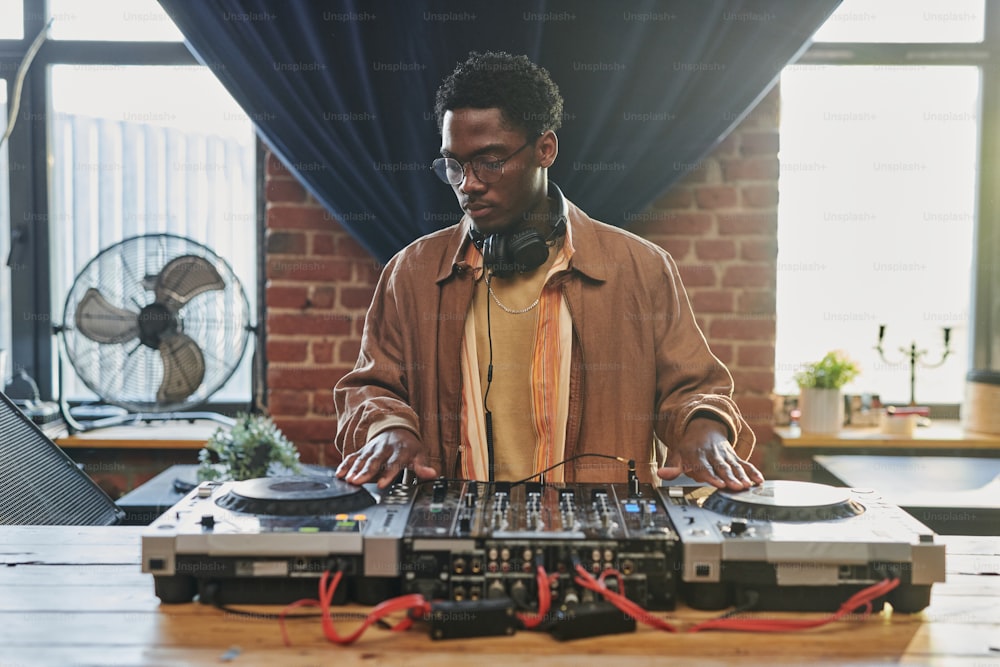 Young black man in casualwear looking at dj board while touching turntables and adjusting musical equipment in studio