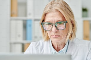 Contemporary mature female online doctor looking at computer screen during medical consultation of patients in office of clinics