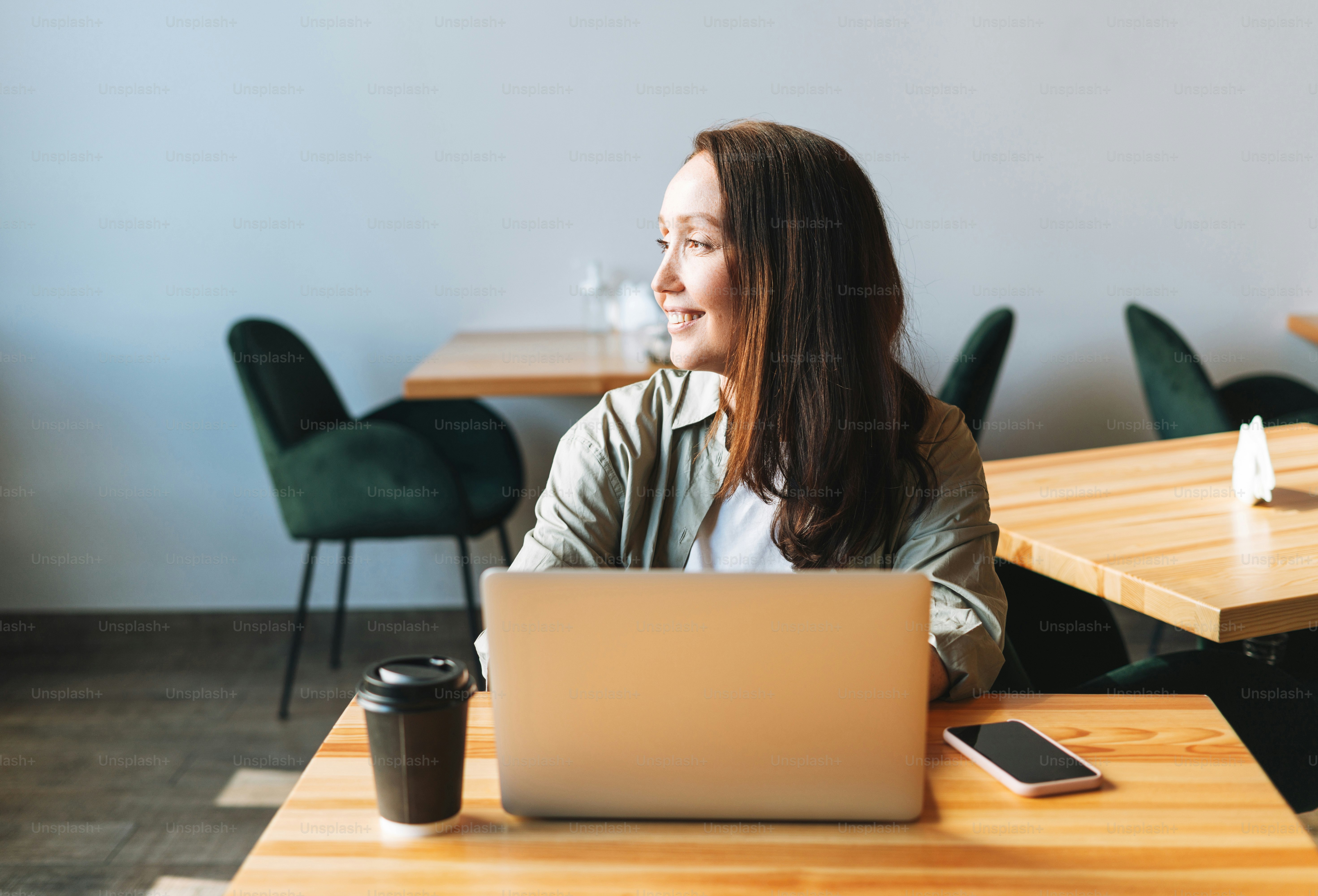 Adult smiling brunette business woman forty years with long hair in stylish shirt working on laptop using mobile phone in cafe