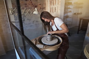 Kunsthandwerkerin sitzt in ihrer Keramikwerkstatt und formt mit einem Werkzeug ein Stück Ton, das sich auf einer Töpferscheibe dreht