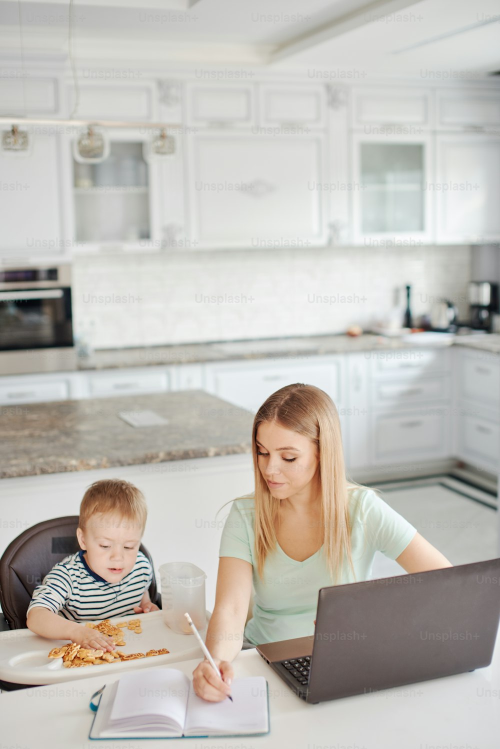 Porträt einer jungen blonden Frau mit Kind zu Hause. Sie benutzt den Laptop, der Sohn sitzt im Kinderstuhl und isst. Zu Hause bleiben, von zu Hause aus arbeiten, beschäftigtes Mutterkonzept