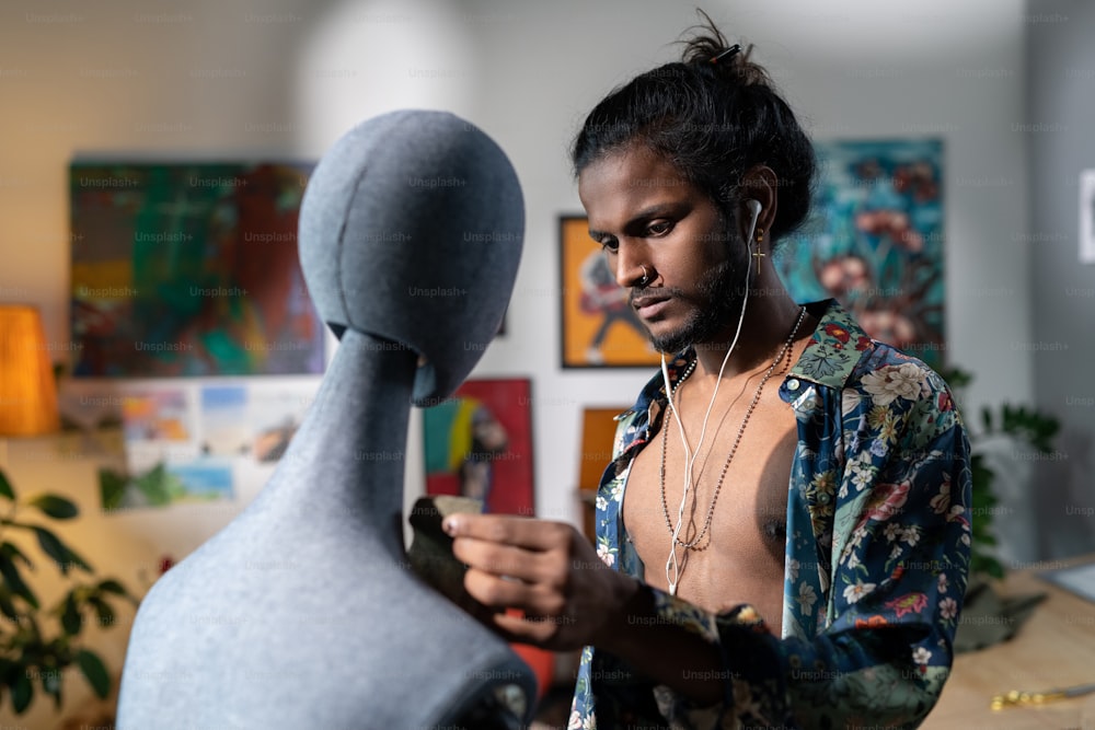 Beau jeune homme indien portant une chemise déboutonnée écoutant de la musique et travaillant sur la conception de vêtements à l’aide d’un mannequin