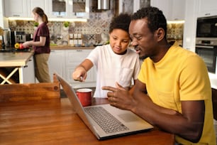 Young African man and cute little biracial boy discussing online video by wooden table in living-room against young female