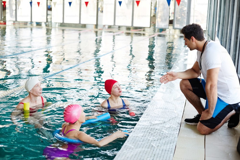 水中で泳ぐ3人のシニアスイマーが、泳ぐ前に若いトレーナーの推薦者と話す