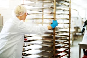 Une jeune travailleuse assez concentrée dans des tissus stériles pousse une étagère en tôle à travers une chaîne de production alimentaire.