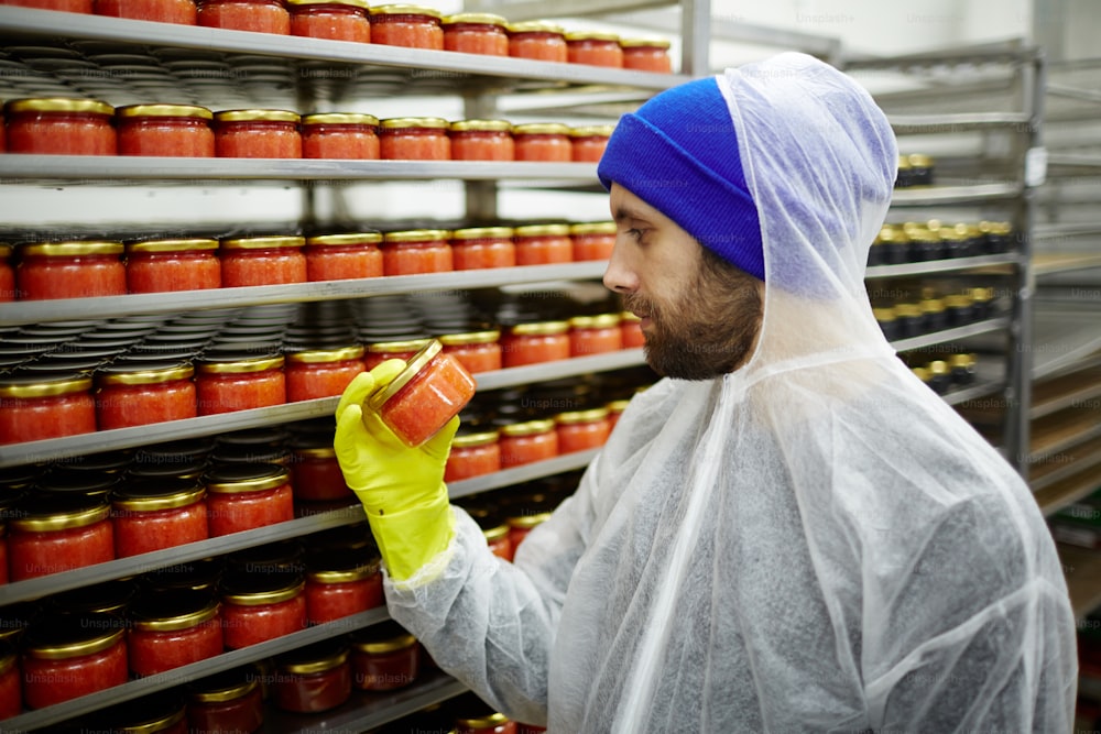 Young man in gloves and protective coveralls examining jar with red caviar after packing