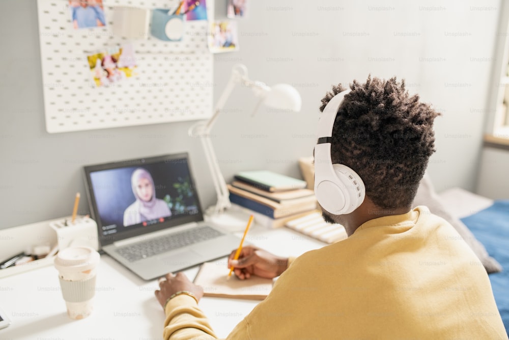 Joven estudiante africano con auriculares sentado frente a la computadora portátil con el profesor en la pantalla y tomando notas durante la lección en línea