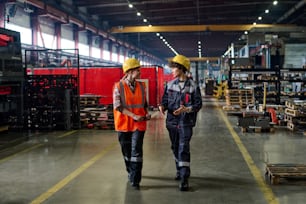 Two workers of modern factory walking down long and wide aisle between industrial equipment and having discussion