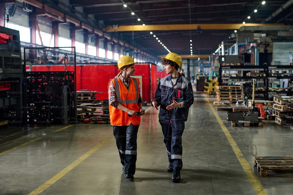 Two workers of modern factory walking down long and wide aisle between industrial equipment and having discussion