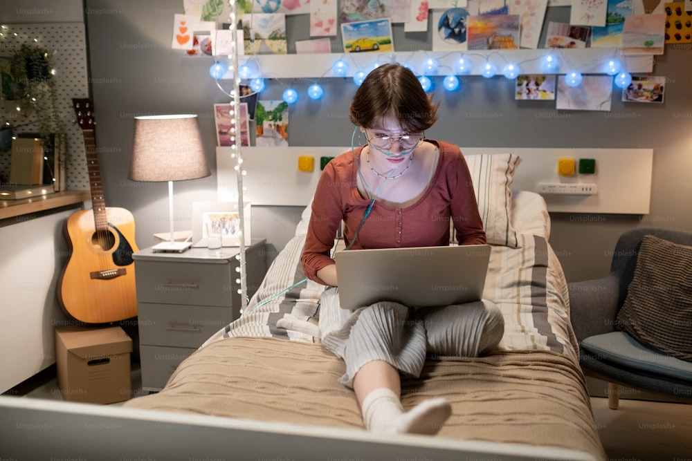 Adolescente seria con computadora portátil en sus rodillas sentada en la cama y trabajando en la red en la sala del hospital